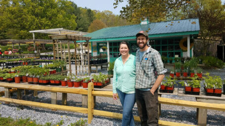 Woman and man in baseball cap standing in front of farmstand with potted plants and a shed in the backfront - Woods Market Garden, Rutland County, Vermont