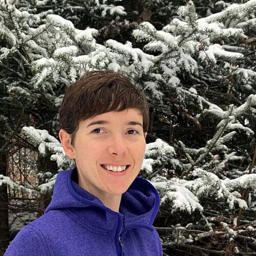 Woman wearing purple jacket smiling at camera with snow covered evergreen tree behind her