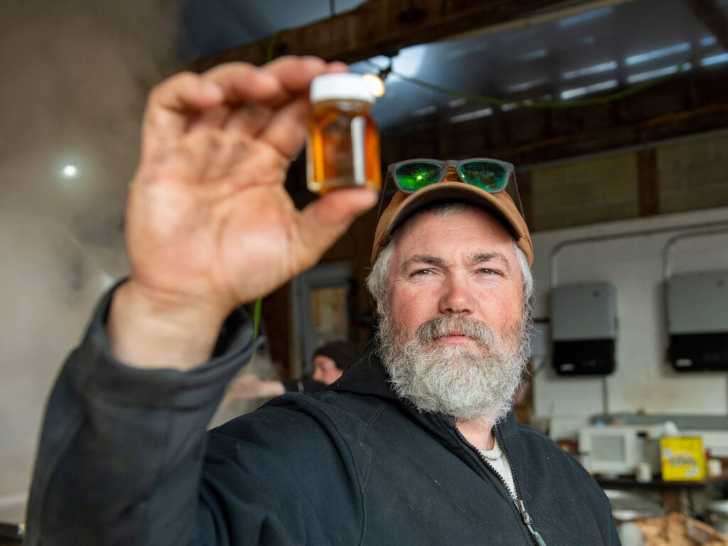 Close-up of man holding small glass jar with golden maple syrup inside - workshed in background. Bunker Farm, Dummerston Vermont