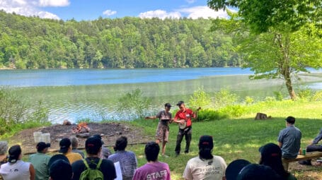 Lakeshore gathering on sunny day with people on picnic benched watching two people play music and sing. Vermont LGBTQ+ camp