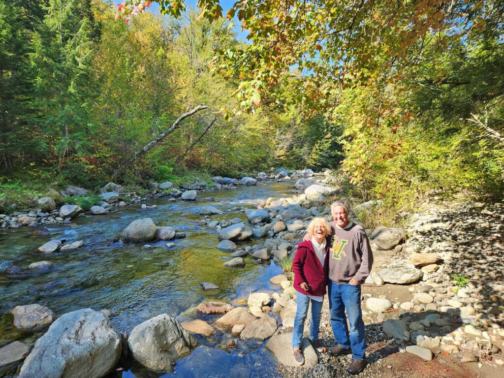 a smiling man and a woman standing at the edge of a river with trees on either side of the water. Don and Sandy Crocker in Montgomery Vermont 