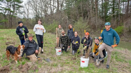 Group of high school students, teachers, and other adults at edge of stream in springtime, with shovels and buckets for planting trees. Shoreham Vermont