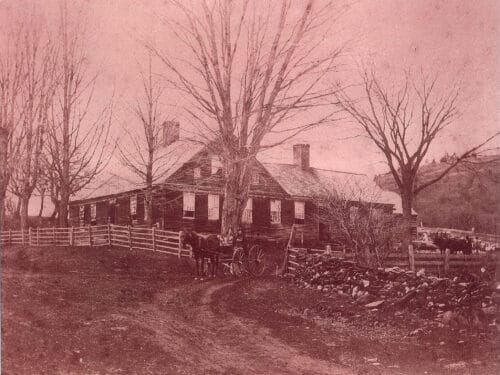 archive photo (sepia toned) of farmhouse with wood fencing and stone walls along boundaries, trees, and two people sitting in horse cart exiting the gate. King Farm Vermont 