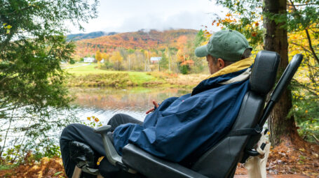 Man sitting with face turned toward pond and forest full of fall colors.