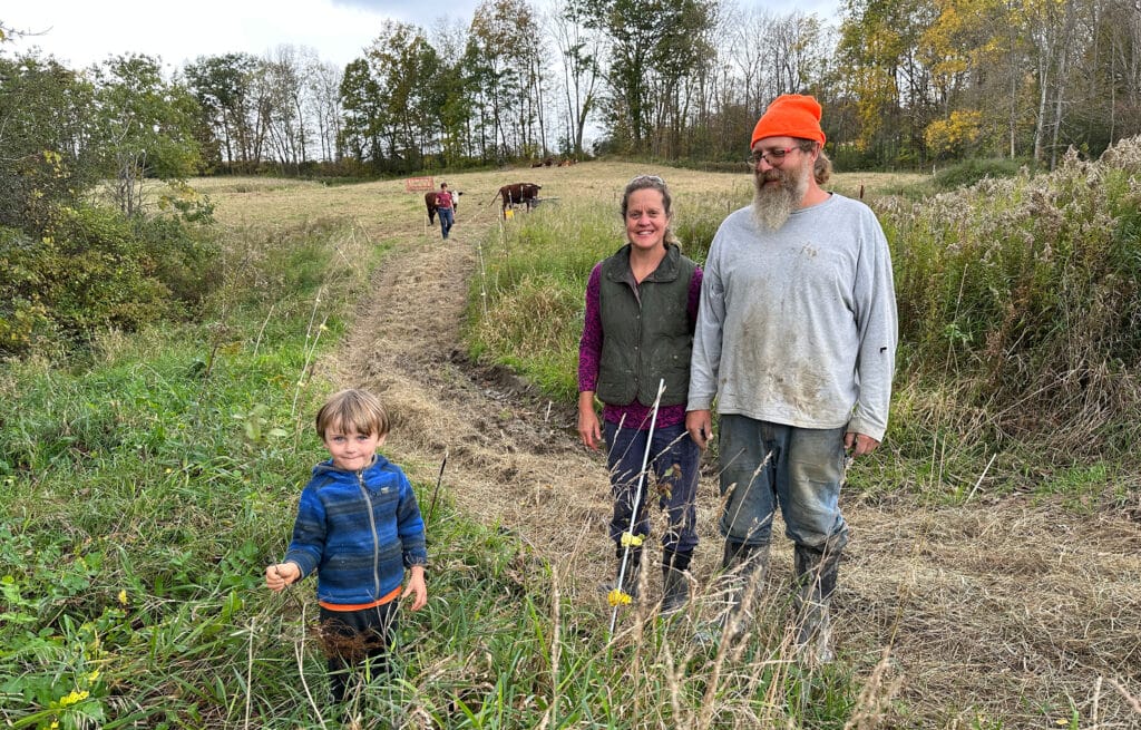 A woman and man standing in a farm field with their small son. West Haven. Soaring Meadow Farm. Vermont