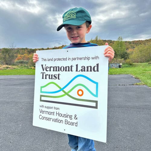 Small boy wearing baseball cap and holding up a sign that reads 'This land protected in partnership with Vermont Land Trust' 