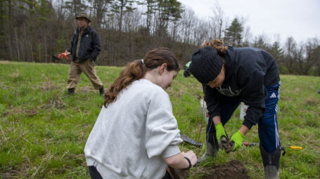 Two students plant a tree in a floodplain field ringed by trees.