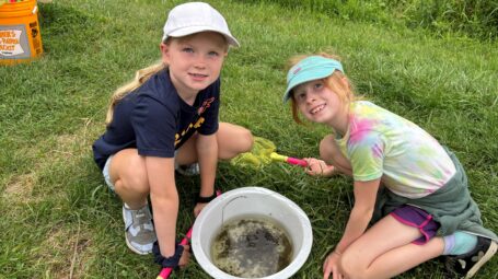 Two smiling girls kneel on grass in front of bucket with water and amphibians.