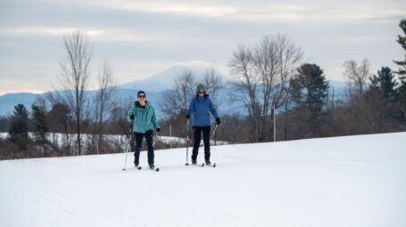 Two cross country skiers ski towards the camera with snowy mountains and trees in the background.