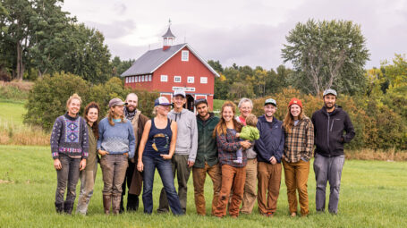 group of smiling men, women, and one toddler standing in a farm field with large red barn behind. The Agrihood Collective, South Burlington, Vermont