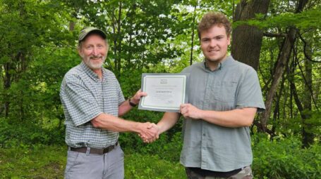 Two men stand holding an award and shaking hands with green trees in the background.