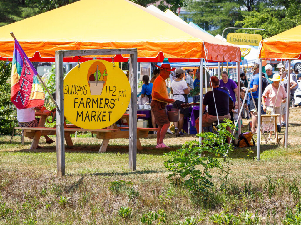 Yellow tent with sign that reads "Sunday 11-2 Farmers' Market" on sunny day with people milling and shopping. Putney Farmers Market Vermont