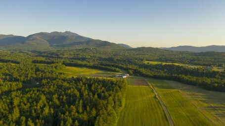 An aerial view of the Brewster Uplands property. Agricultural fields and forest are in the foreground with the Mount Mansfield ridge behind.