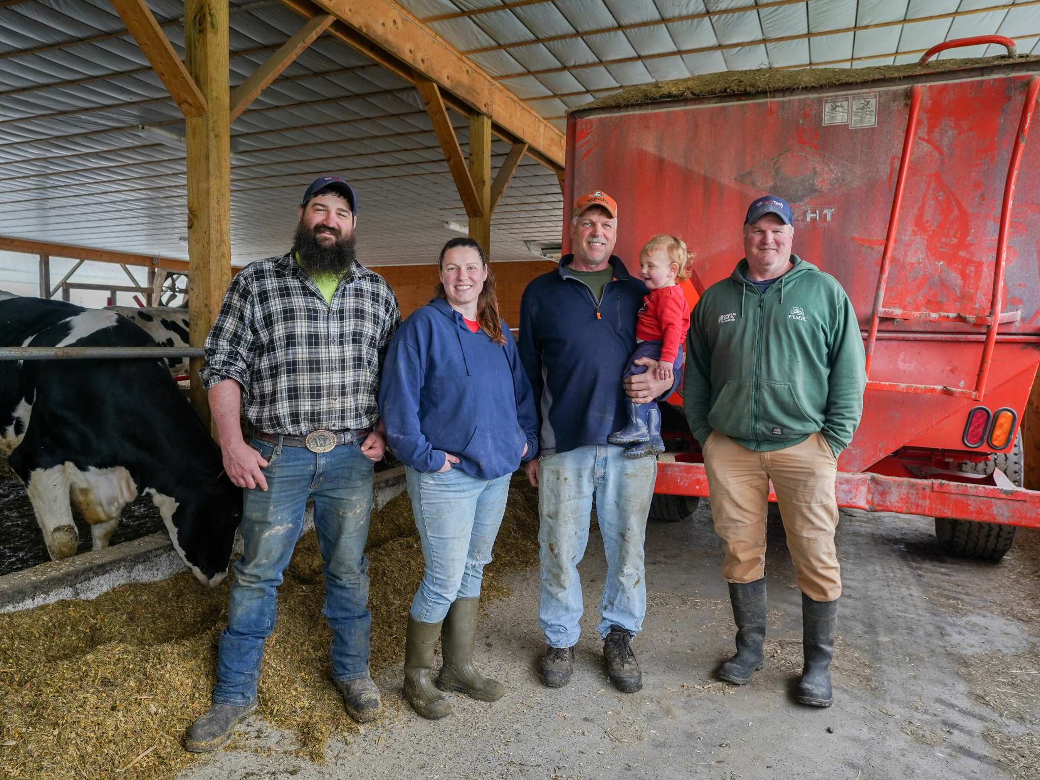 Four adults and one baby stand in a barn with cows in the background.