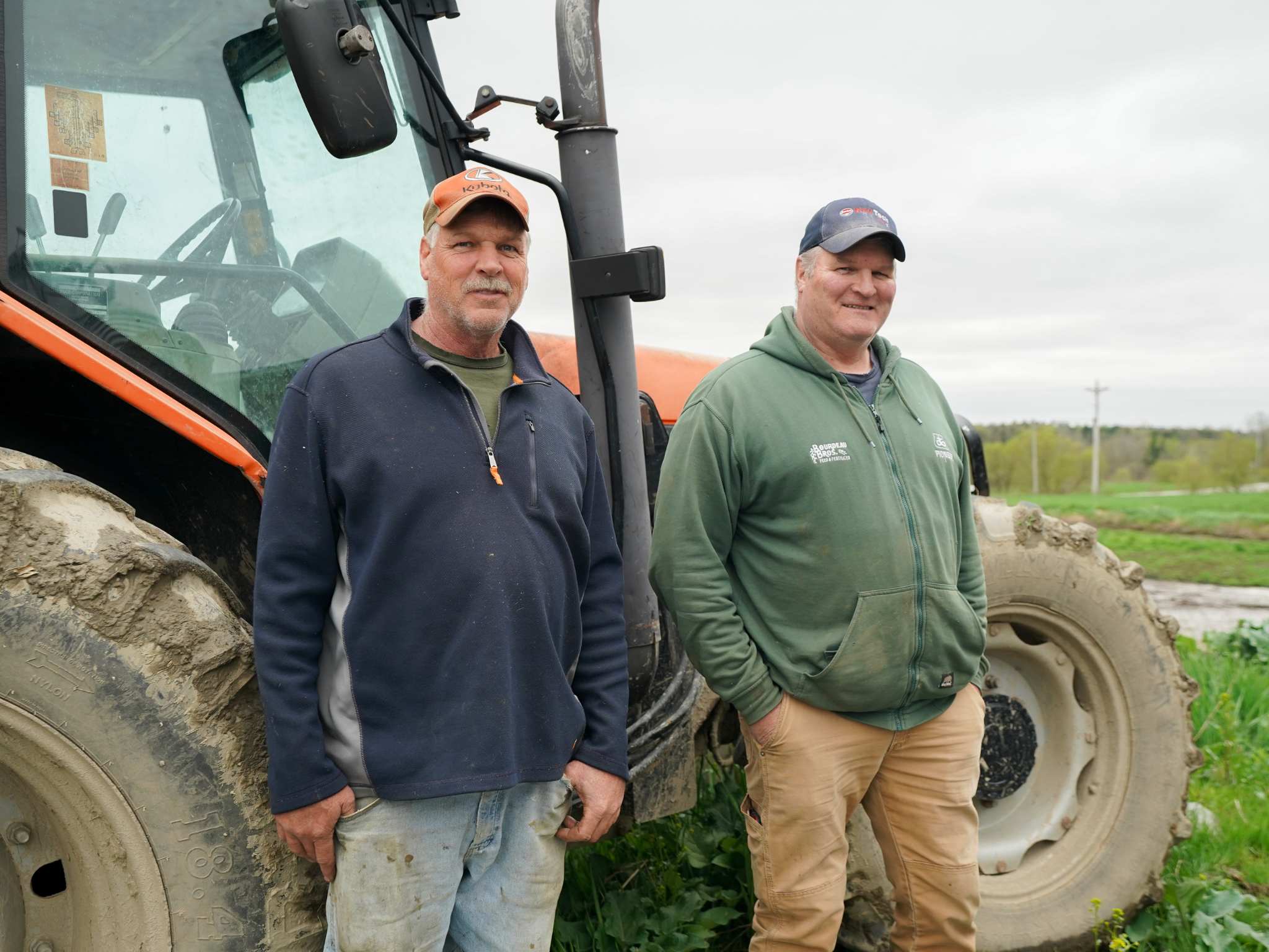 Two older men wearing hats stand smiling in front of a tractor