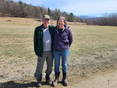 A couple stands together on a dirt road with a field and forest in the background.
