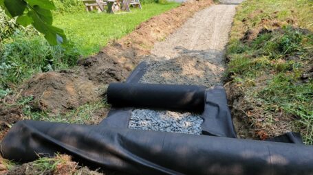 A trail build in progress shows the cross-section or gravel and fabric layers running through disturbed ground into the woods, with grass linking the sides.
