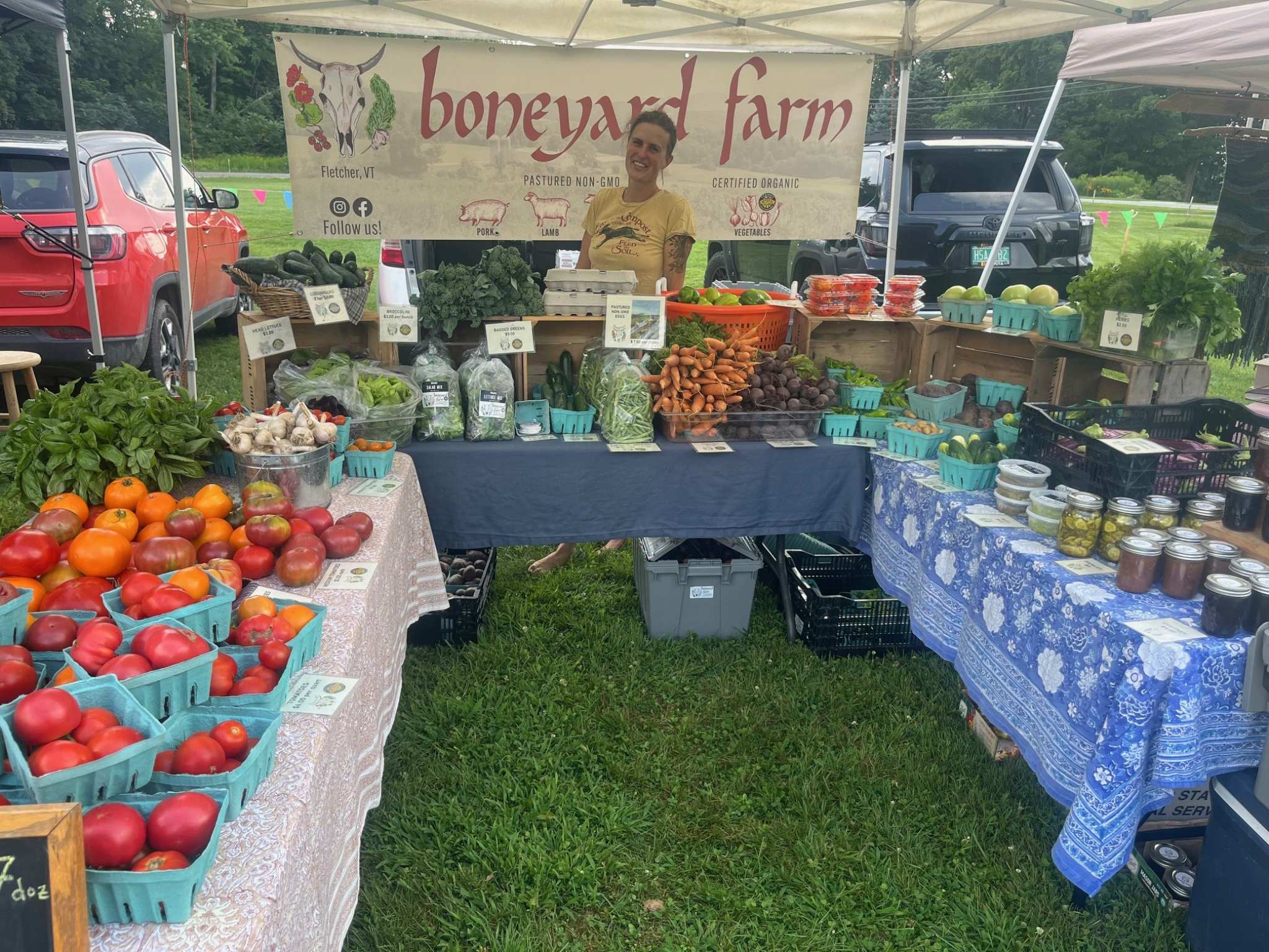 A woman stands in front of a sign that reads "boneyard farm" and behind tables loaded with produce.