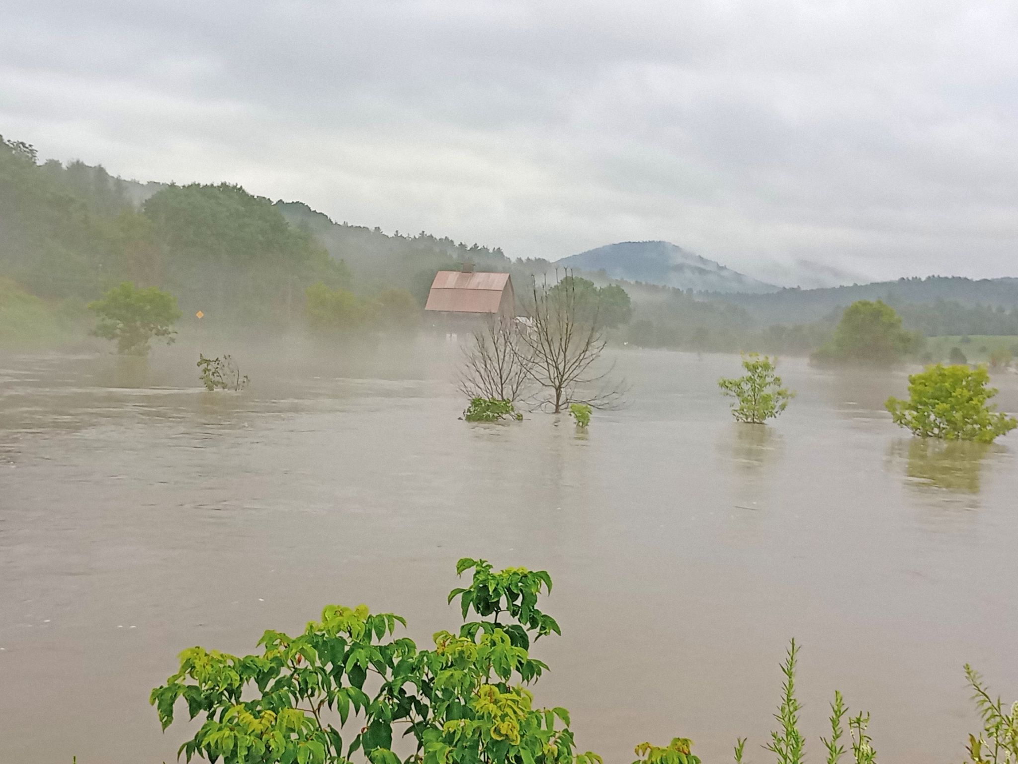 Mist rises from muddy flooded water with old farm in the background.