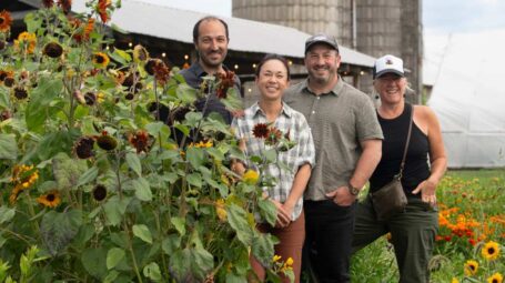 Four people stand partially behind an array of sunflowers, smiling at the camera.