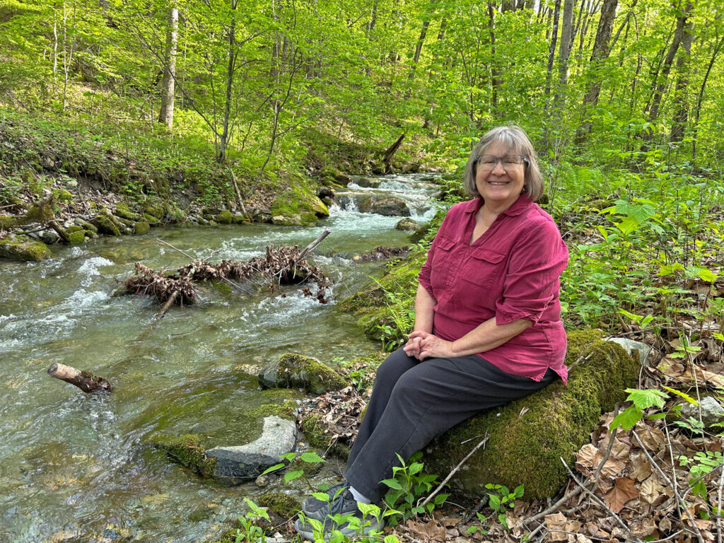 Woman seated on rocks by a wooded stream, smiling at camera. Arlington