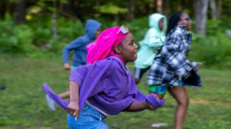 A black girl with a pink headdress and purple sweater runs with other kids through a field