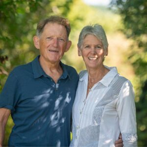 A smiling older white couple with greenery in the background