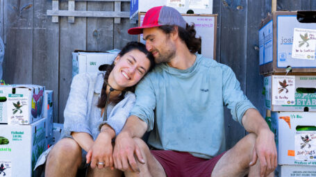 A smiling woman in braids leans her head on a man's shoulder. He wears a cap and rests his head on hers.