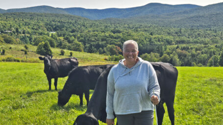 An older woman stands in a field with cattle behind her and mountains in the background