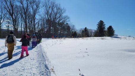 A large group of people snowshoe through a snowy field, with trees in the background.