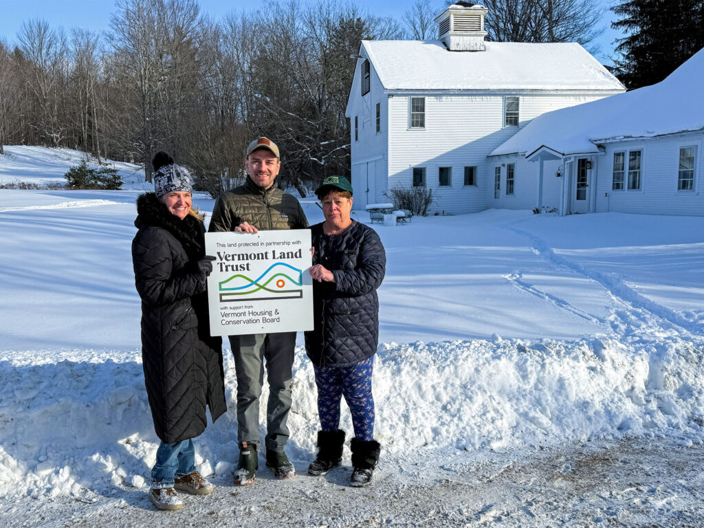 Three people standing holding sign that says 'this land protected in partnership with Vermont Land Trust' by side of road with white farmhouse and deep snow behind