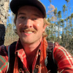 close up photo of young man wearing baseball cap and red plaid shirt with pine woods behind. Vermont