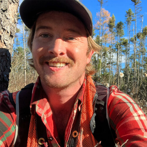 close up photo of young man wearing baseball cap and red plaid shirt with pine woods behind. Vermont