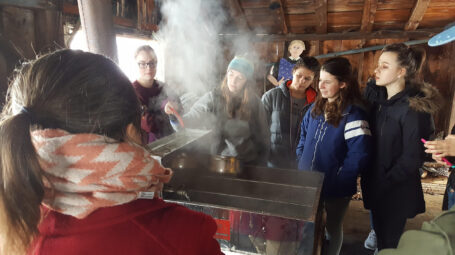 Students and a teacher stand around a steaming maple arch.