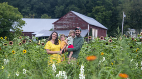A man and a woman stand in a farm field, each holding a child.