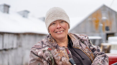 woman farmer wearing wool hat and seated on tractor smiles at camera. Bakersfield Vermont