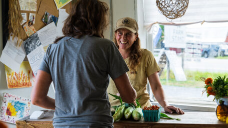 A farmer at a farmstand smiles at a customer.