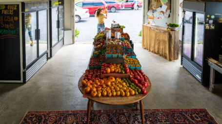 An interior of a farm stand with a table full of vegetables, flanked by reach-in coolers.