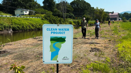 Sign reading 'Clean Water Project' and showing map of Vermont, with people in distance of newly planted area along river. Marshfield Vermont