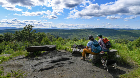 Two hikers and a dog sit on mountain-top benches overlooking a green valley.