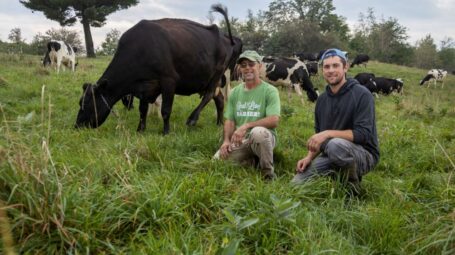 Two farmers kneel in a green pasture next to a cow.