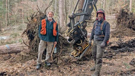 Two workmen wearing safety gear at a tree management site with large tractor and exposed tree rootwad. Vermont