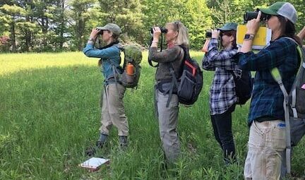 A group of women stand in a clearing, looking through binoculars.