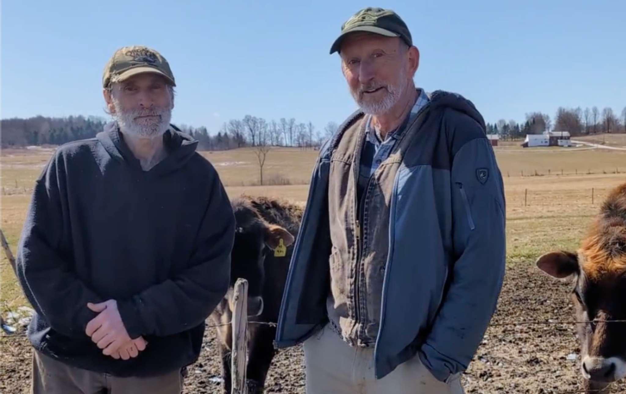 Two men stand together with cows in a pasture behind them.