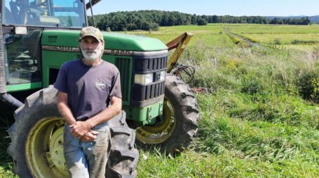A man stands in front of a green tractor with farm fields and forest in the background.