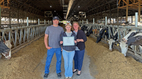 Three people stand in the center aisle of a cow barn, an older man on the left, a young woman holding a certificate in the middle, and an older woman on the right.