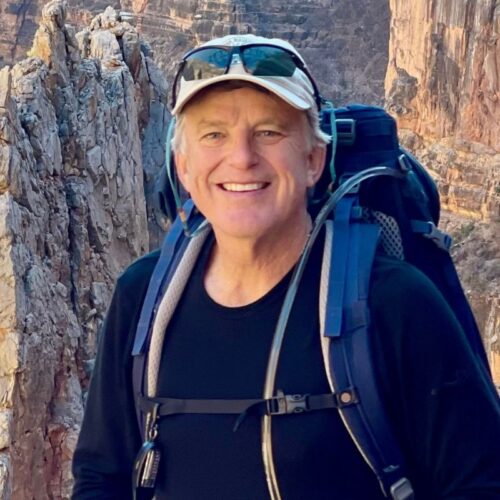 A smiling white man wearing a hat and a backpack with rocky cliffs behind him.