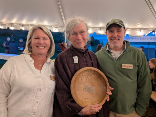 Three people smiling, the center one holding a gifted wooden bowl.