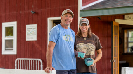 farm couple standing outside red barn and farmstand